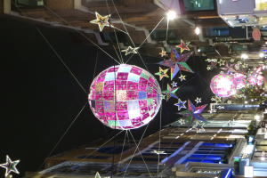 Eine Stadtstraße bei Nacht voller Lichter und Dekorationen, mit einem großen Diskokugel an einem Gebäude hängend, mit beleuchteten Gebäuden, die eine festliche Atmosphäre schaffen.