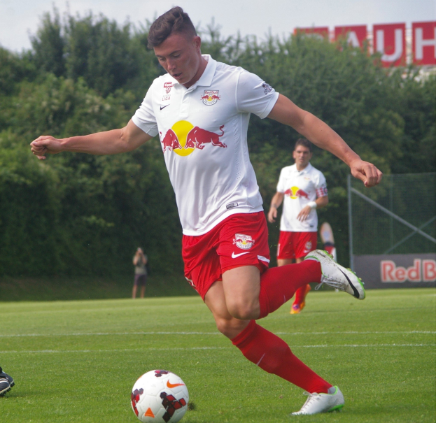 Ein professioneller Fußballspieler, der einen Ball auf einem Rasenfeld mit Bäumen und einem klaren blauen Himmel im Hintergrund kickt, in der Nähe einer Tafel mit der Aufschrift "RB Leipzig."