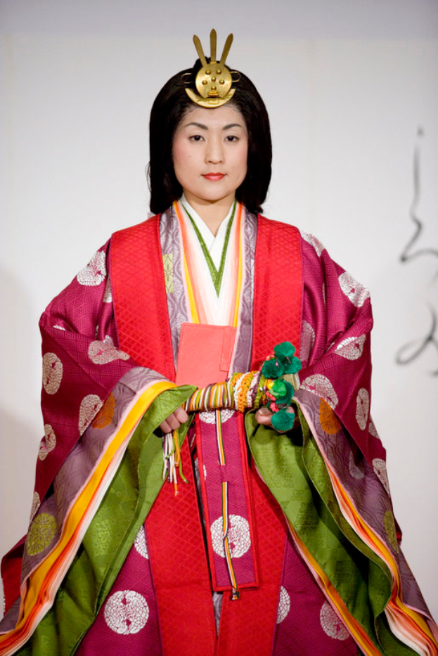 Eine Frau in einem traditionellen japanischen Kimono, das ein leuchtendes rotes und grünes Kleid mit aufwändigen Mustern trägt, steht selbstbewusst auf einem Laufsteg, ihr Haar ordentlich zu einem Knoten gebunden.