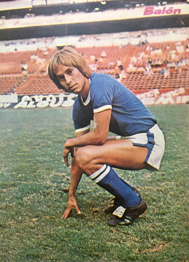 Ein Mann kniet auf einem Fußballfeld umgeben von Menschen auf Stühlen und Schildern mit Text, trägt Schuhe.