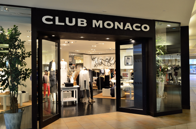 Ein Club Monaco-Laden in einem Einkaufszentrum mit Glaswänden und -türen, Schaufensterpuppen in Kleidern, Auslagetischen, einem Topf mit einer Pflanze und Deckenleuchten.