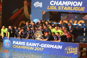 Eine Gruppe von Paris Saint-Germain-Spielern in Medaillen feiert mit einem Pokal vor einer Tafel mit Text und Logos, mit einem Netz auf der rechten Seite und einem Bildschirm im Hintergrund.