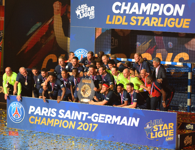 Eine Gruppe von Paris Saint-Germain-Spielern in Medaillen feiert mit einem Pokal vor einer Tafel mit Text und Logos, mit einem Netz auf der rechten Seite und einem Bildschirm im Hintergrund.