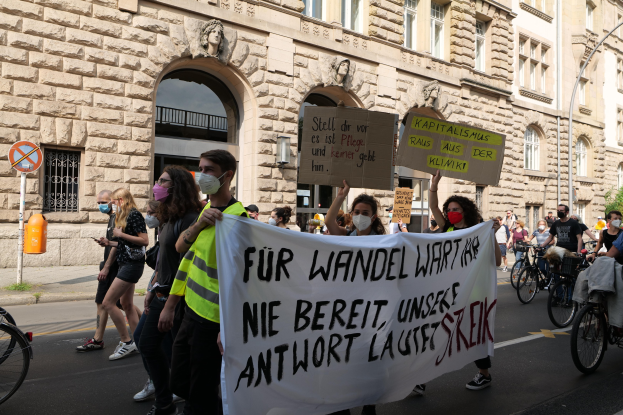 Eine Gruppe von Menschen marschiert bei einer Protestaktion gegen die Anti-Kriegsbewegung in Berlin, einige halten Schilder und andere fahren Fahrräder, mit einem Gebäude im Hintergrund, das Fenster, Bögen, Säulen und Skulpturen aufweist.