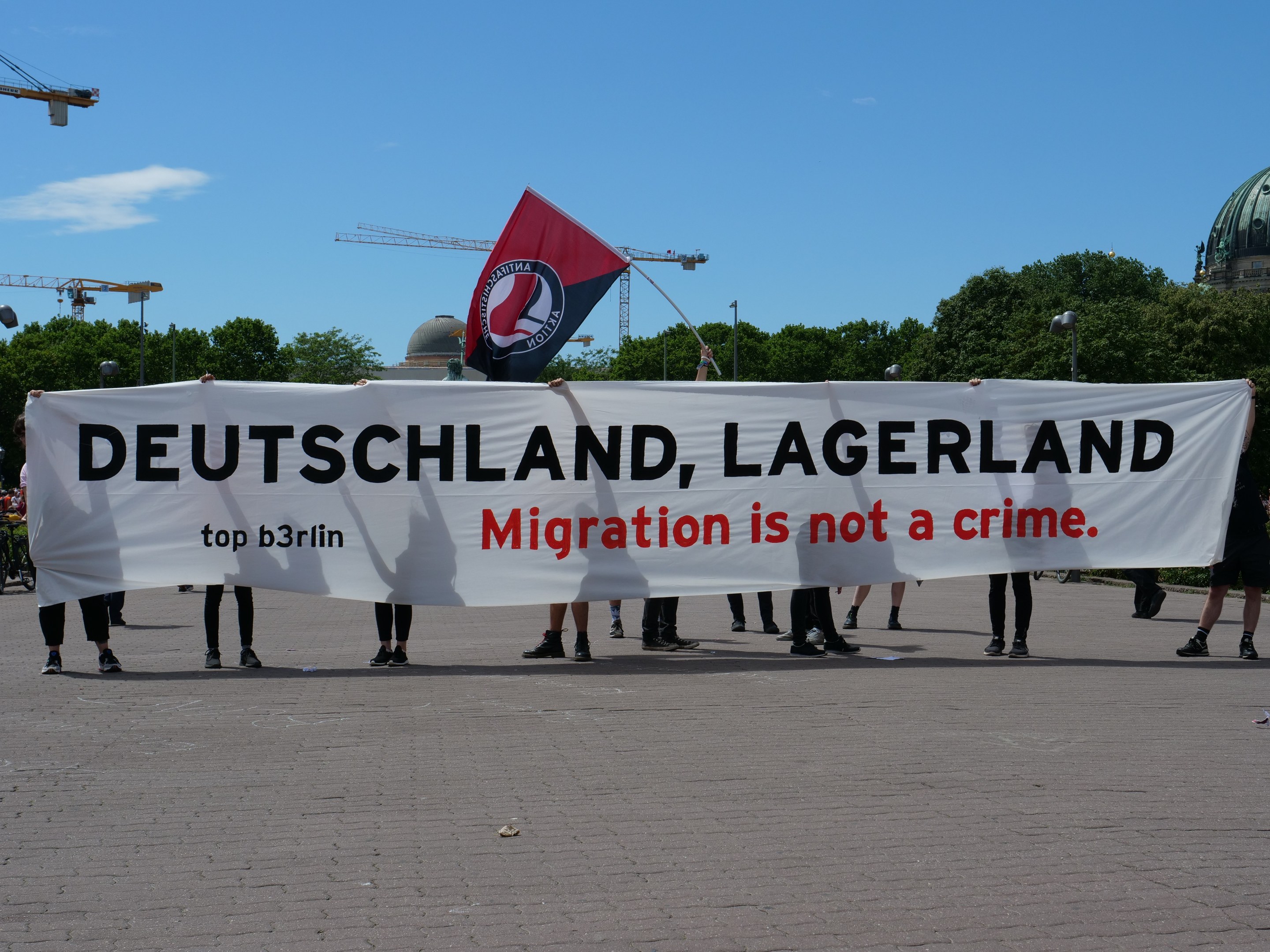 Eine Gruppe von Menschen hält ein Transparent mit der Aufschrift "Deutschland, Lagerland Migration ist kein Verbrechen" und eine Flagge, mit Straßeninfrastruktur und Gebäuden im Hintergrund bei bewölktem Himmel.