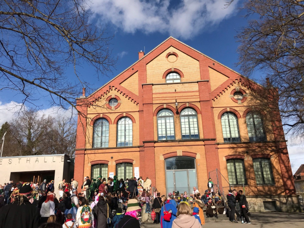 Große Gruppe von Menschen, die vor einem mehrstöckigen Gebäude mit Bäumen und einem bewölkten Himmel im Hintergrund stehen, einige tragen Mützen und tragen Taschen.