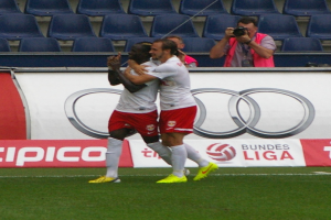 Zwei Fußballspieler feiern ein Tor auf einem Feld, umgeben von einer Gruppe von Menschen, mit einer Person, die eine Kamera hält, einem Banner mit Text, einigen Stühlen und einer Wand im Hintergrund.