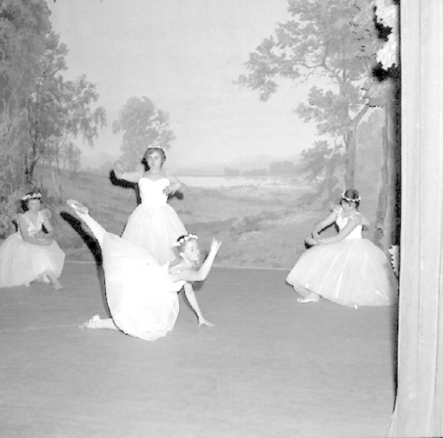 Vier junge Balletttänzerinnen in weißen Kleidern tanzen auf einer Bühne mit einer gemalten Kulisse aus Bäumen und Himmel in Schwarz-Weiß.