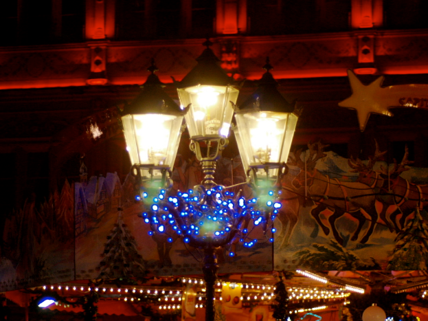 Eine Straßenlaterne in einer nächtlichen Stadt, geschmückt mit Lichtern und Dekorationen, steht vor einem Gebäude und verbreitet eine festliche Stimmung.