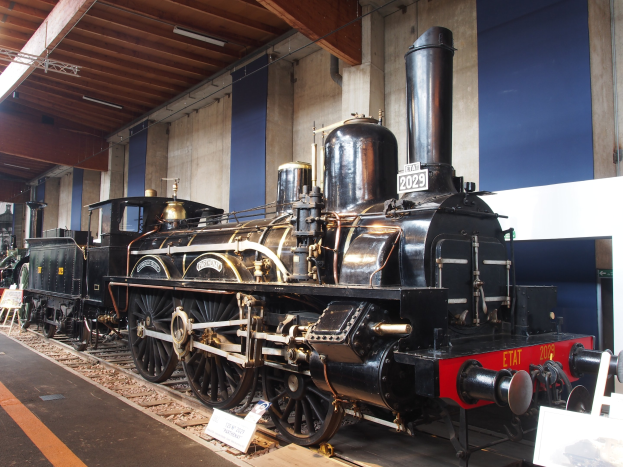 Alte Dampflokomotive in einem Museums Setting mit umliegenden Eisenbahnschienen, Informationsschildern und Beleuchtungsk├Ârpern an einer Wand und Decke.