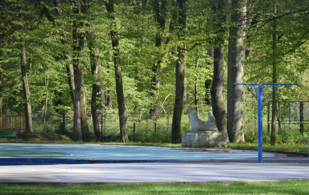 Ein Basketballplatz in einem Park umgeben von Bäumen, mit einer Bank links, einem Zaun im Hintergrund und Gras unten.