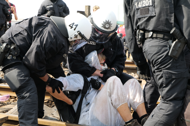 Eine Gruppe von Polizisten in schwarzer Einsatzkleidung verhaftet einen Mann in einem weißen Kleid auf einem Bahngleis, mit anderen Menschen im Hintergrund.