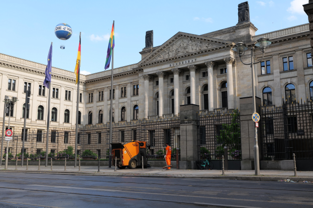 Großes modernes Regierungsgebäude mit vielen Fenstern, umgeben von Straßeninfrastruktur, Fahrzeugen, Fußgängern, Flaggen und Bäumen unter einem bewölkten Himmel, identifiziert als der Bundestag in Berlin, Deutschland.