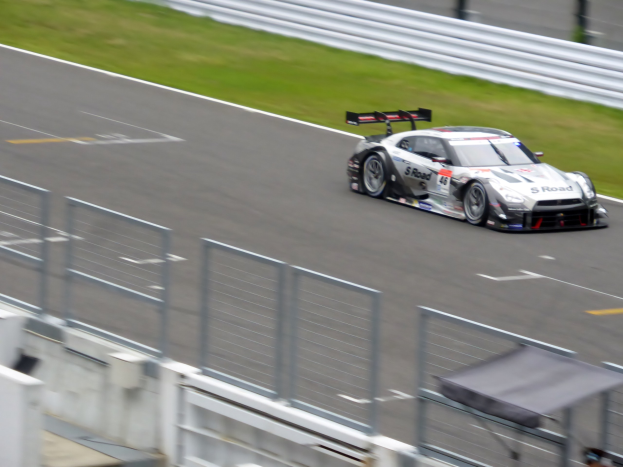 Ein Nissan GT-R Nismo GT3 Rennwagen auf der Nürburgring 24 Stunden Strecke, mit einem Zaun und Gras auf beiden Seiten, einem Schuppen und ein paar Leuten unten und einem unscharfen Hintergrund.