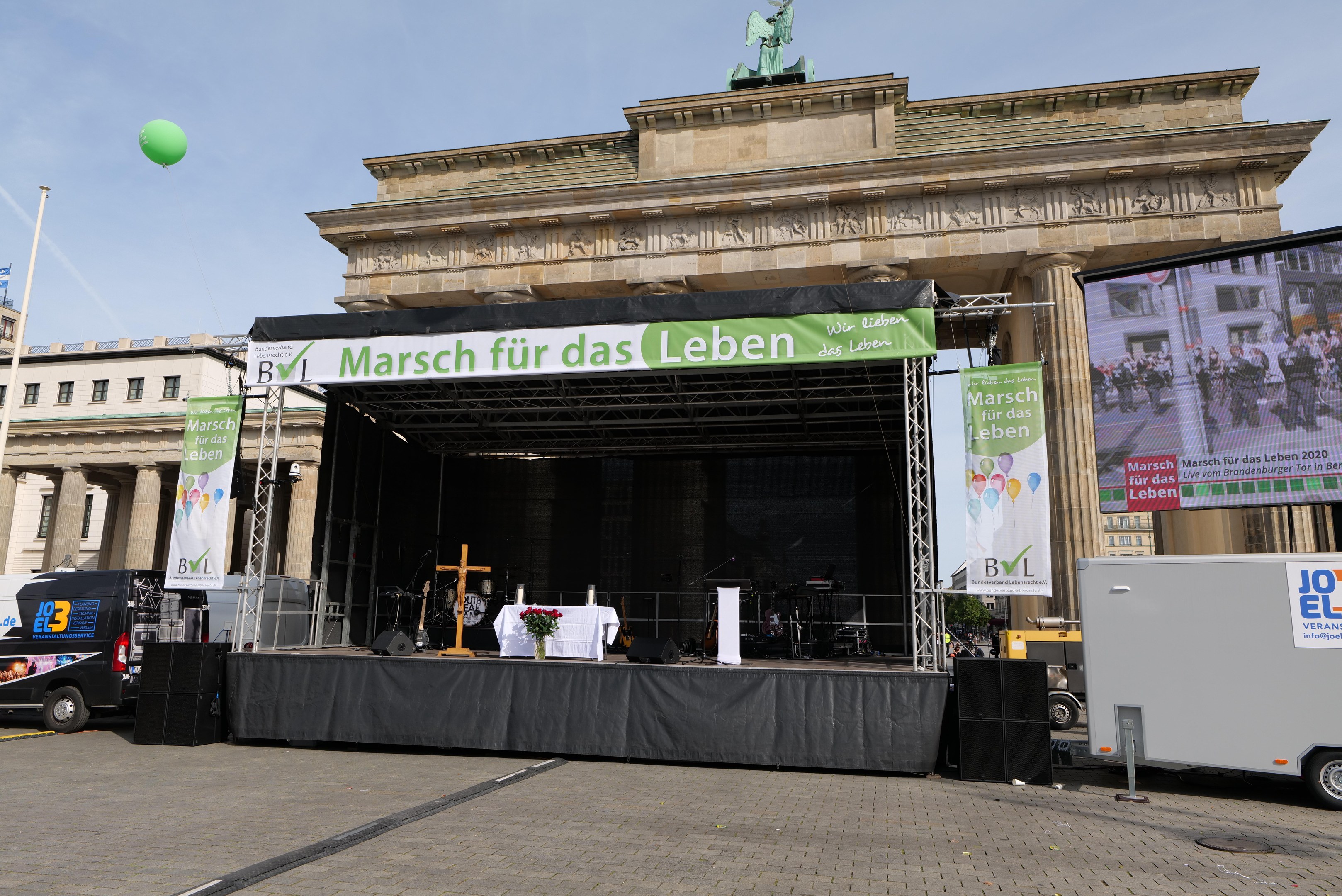 Bühne vor dem Brandenburger Tor mit einem Tisch, Bannern, Lautsprechern, Fahrzeugen, Gebäuden, einer Statue, einer Flagge und Wolken.
