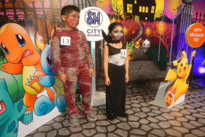 Zwei Kinder in Halloween-Kostümen stehen nebeneinander vor einer Wand mit einem Gemälde eines Gebäudes, Bäumen, Laternenmasten und einem Zaun, mit Schildern, die Text und Tierbilder zeigen, und festlicher Beleuchtung.