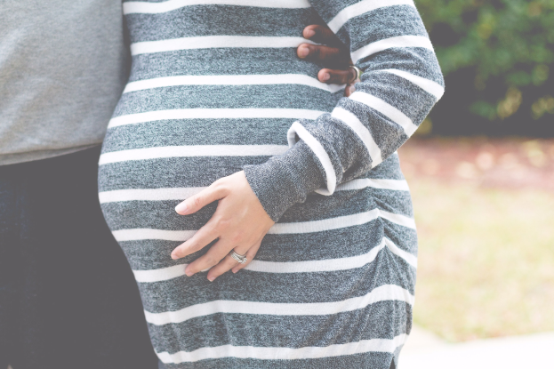 Schwangere Paare in Schwangerschaftskleidung, die zusammen mit den Händen auf dem Bauch stehen, umgeben von Bäumen.