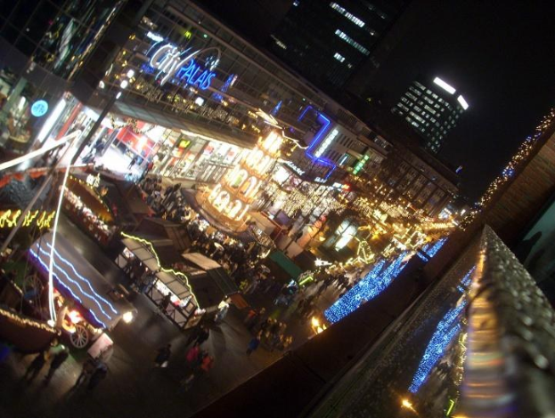 Eine belebte Stadtstraße bei Nacht voller Menschen, die gehen, beleuchtet von Gebäuden und Marktständen, mit dunklem Himmel im Hintergrund.