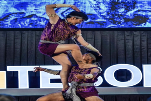 Zwei Akrobaten führen auf der Bühne eine Nummer auf, wobei der Mann die Frau in seinen Armen hält und beide purpurne Kleider tragen, während im Hintergrund eine Menschenmenge und ein Bildschirm zu sehen sind.