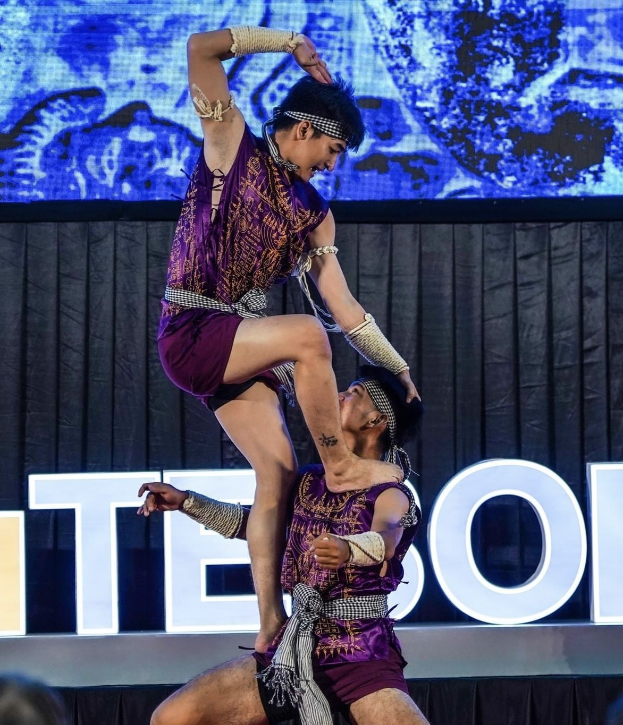 Zwei Akrobaten führen auf der Bühne eine Nummer auf, wobei der Mann die Frau in seinen Armen hält und beide purpurne Kleider tragen, während im Hintergrund eine Menschenmenge und ein Bildschirm zu sehen sind.