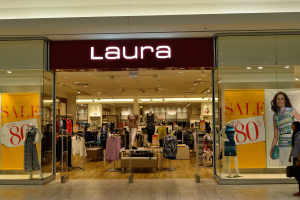 Ein Laura-Kleidungsgeschäft in einem Einkaufszentrum mit Schaufensterpuppen in Kleidern, hängenden Kleidern, Informationsschildern, Deckenleuchten und einer Person, die eine Tasche trägt.