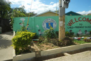 Eine grüne Wand mit einem Schild, das "Willkommen in der kinderfreundlichen Schule" trägt, umgeben von Häusern, Bäumen, Pflanzen und einem Pfahl mit einer Lampe.