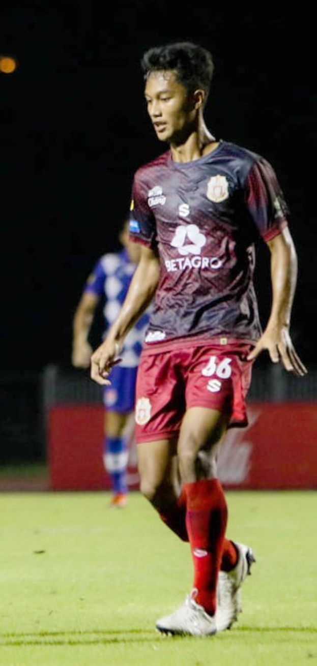 Ein Mann in einem rotbraunen Hemd und roten Shorts, der auf einem Feld Fußball spielt, mit einer anderen Person im Hintergrund und einer Tafel mit Schrift darauf.