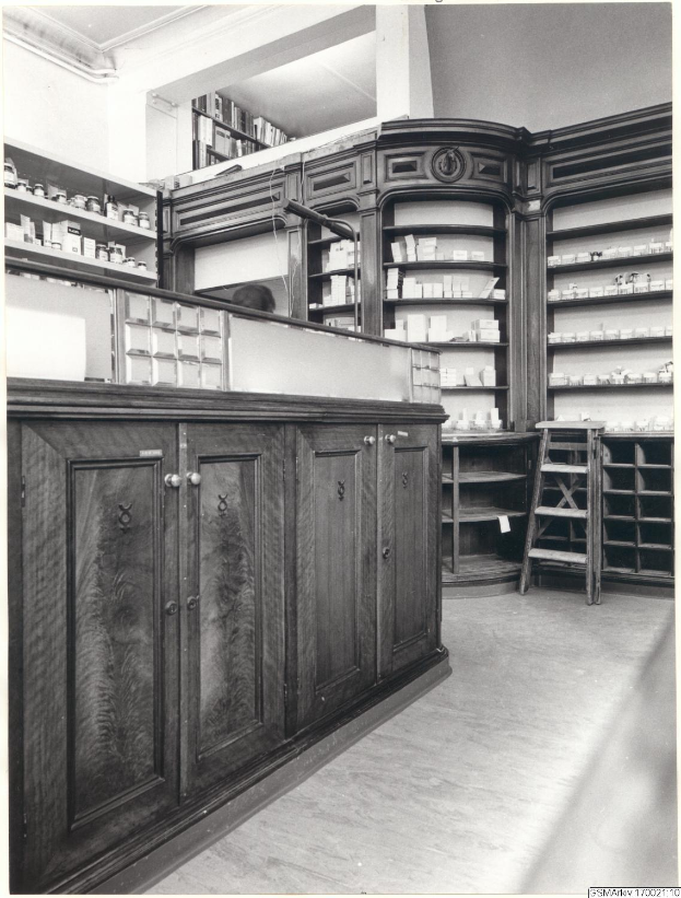 Ein Schwarz-Weiß-Foto eines Apothekeninneren mit einer Theke, Schränken, einer Leiter und Regalen, die mit verschiedenen Gegenständen gefüllt sind, mit Text oben.