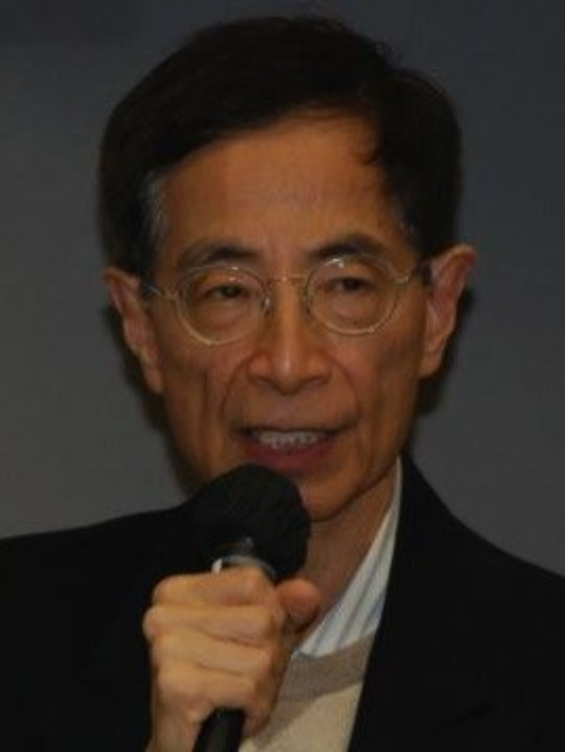 Ein Mann in einem schwarzen Blazer und Brille hält ein Mikrofon mit ernster Miene, er scheint ein Ökonom zu sein.