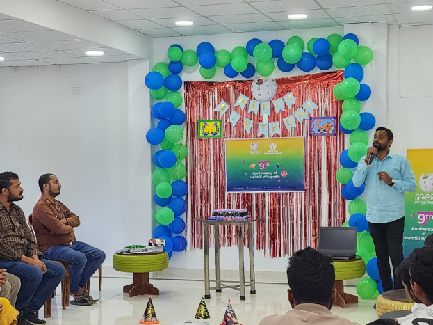 Eine Gruppe von Menschen, die auf Stühlen sitzen und einer dekorierten Bühne mit Ballons, einem Banner, einer Uhr und anderen Gegenständen zugewandt sind, mit einem Tisch mit einem Laptop und einer Person, die ein Mikrofon hält, während einer 9. Geburtstagsfeier.