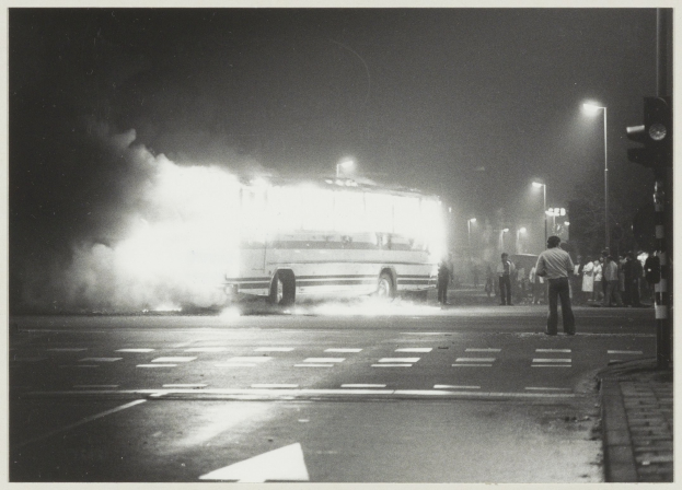 Schwarzes und weißes Foto eines brennenden Busses am Straßenrand mit einer Gruppe von Menschen auf dem Gehweg rechts und beleuchteten Straßenlaternen im Hintergrund.