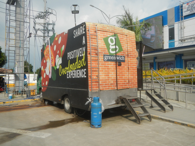 Ein Lkw parkt vor einem Gebäude mit einer Seite, auf der eine Tafel mit der Aufschrift "Teilen Sie ein positiv überladenes Erlebnis" zu sehen ist, mit einer Treppe, einem Zylinder auf dem Boden, Geländern, Strommasten mit Drähten, Laternen, Bäumen und einem bewölkten Himmel im Hintergrund.