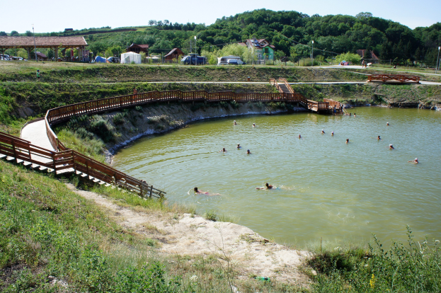 Gruppe von Menschen, die in einem Gewässer umgeben von Grünfläche, einer Brücke, Treppen, Schuppen, Fahrzeugen, Pfählen und einem klaren blauen Himmel schwimmen.