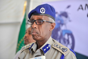 Ein Mann in Polizeiuniform mit Mütze und Brille steht an einem Podium mit Mikrofonen, daneben eine weitere Person, eine Fahne und ein Banner.