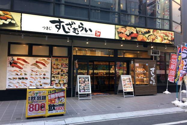 Außenansicht einer Sushi-Bar in Tokyo mit Glaswänden, Essensauslagen, Schildern und einer Straße davor.