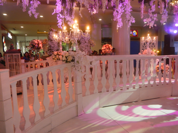 Eine Hochzeitsfeier mit einem weißen Geländer, das mit Blumen geschmückt ist, Kronleuchtern an der Decke, Menschen, die sich darum versammeln, Säulen und Lichter im Hintergrund und ein Bildschirm auf der rechten Seite.