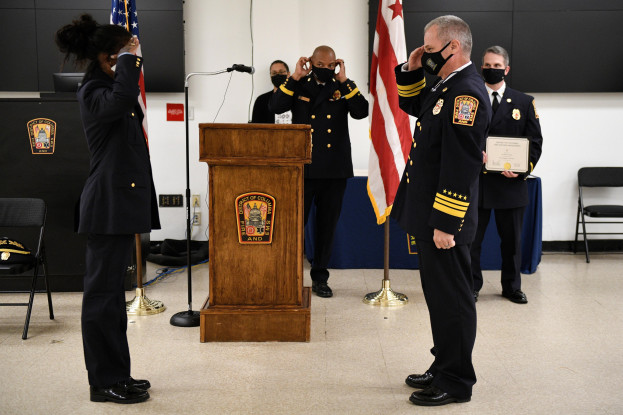 Gruppe uniformierter Personen in Masken vor einem Podium mit Mikrofon, das eine Urkunde während einer Vereidigung eines neuen Feuerwehrchefs hält, umgeben von Fahnen, Stühlen, Tischen und einer Wand.