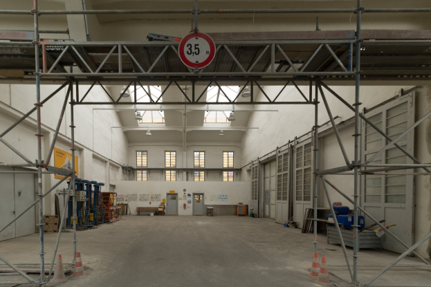 Großes Lagerhaus-Innenraum mit ausgedehnten Gerüsten, Verkehrskegeln, Metallstangen, einem Schild, Maschinen, einer Tür, Fenstern, Wänden, Deckenlampen und einem Dach.