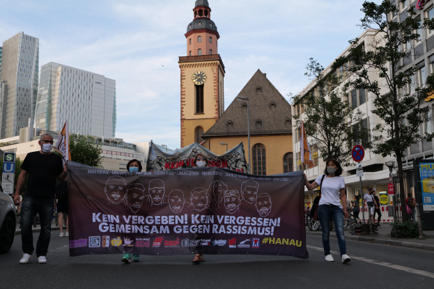Eine Gruppe von Menschen in Masken, die eine Straße entlanggehen und ein Banner halten, mit einem geparkten Auto links, Gebäuden, Bäumen, Schildern, Polen, einem Uhrenturm und einem klaren blauen Himmel im Hintergrund.