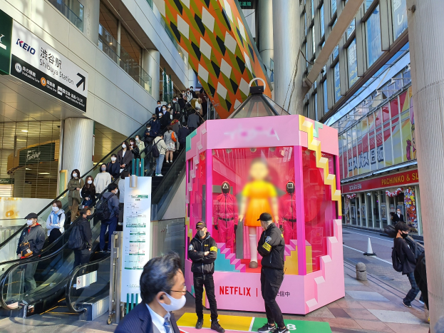 Eine Gruppe von Menschen steht vor einem gläsernen Gebäude mit Säulen und Texttafeln, einige tragen Masken, während eine Rolltreppe mit Menschen zu sehen ist; eine pinkfarbene Box in der Mitte zeigt Schaufensterpuppen und trägt den Text "Netflix's new Netflix booth in Tokyo."
