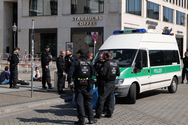 Polizeibeamte vor einem Starbucks-Café mit einem Van auf der rechten Seite und Menschen auf der linken Seite, mit einem Gebäude, einem Schild, einem Lichtmast und einem Zaun im Hintergrund.