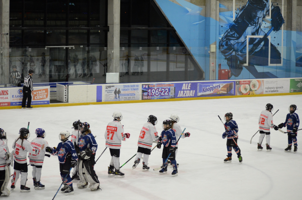 Gruppe junger Menschen beim Eisschnelllaufen auf einer Indoor-Eisbahn, die Helme tragen und Stöcke halten, mit einer Person auf der linken Seite und Bannern, Säulen und Glaswänden im Hintergrund.