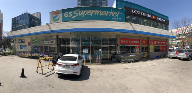 Ein Auto vor einem "GS Supermarkt"-Laden mit anderen Fahrzeugen, Verkehrskegeln und verstreuten Gegenständen auf der Straße; Gebäude, Bäume und ein klarer blauer Himmel im Hintergrund.