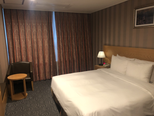 Hotelzimmer mit einem großen Bett, Stuhl, Tisch, Telefon, Lampe, gerahmtes Foto an der Wand, Fenster mit Vorhängen und Deckenbeleuchtung.