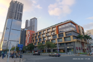 Eine belebte Stadtstraße mit hohen Gebäuden im Hintergrund, Fahrzeugen auf der Straße, Fußgängern auf dem Gehweg und Bäumen, die die Straße säumen, unter einem bewölkten Himmel, mit einem Bürogebäude mit grüner Dachterrasse im Vordergrund.