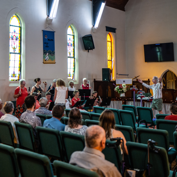 Eine Gruppe von Menschen sitzt auf Stühlen in einer Kirche, ein Mann steht vorne mit einem Mikrofon in der Hand, umgeben von Musikinstrumenten, einem Tisch mit einer Blumenvase, einem Lautsprecher, einer Fahne mit Text, Fenstern und Deckenleuchten.