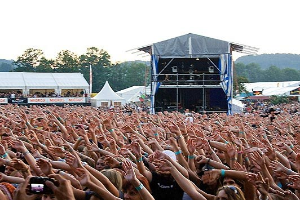 Eine große Menschenmenge auf einem Musikfestival mit erhobenen Händen, einige halten Telefone, vor einer Bühne mit Künstlern, weißen Zelten, Bäumen und einem klaren blauen Himmel.