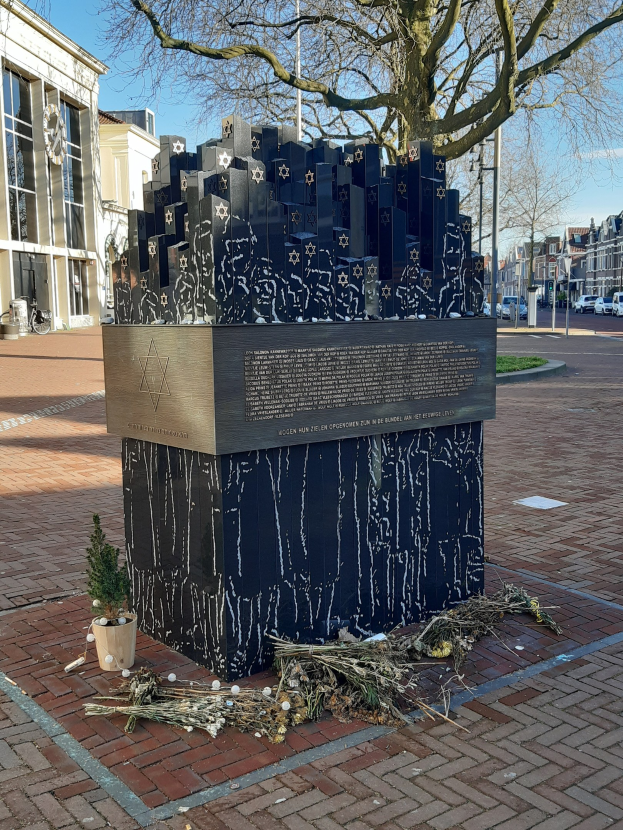 Denkmal in einem Stadtplatz für Holocaust-Opfer mit einem Baum davor, einem Topfgewächshaus auf dem Boden, verstreuten trockenen Blättern und Gebäuden, Fahrzeugen, Masten und Bäumen im Hintergrund.