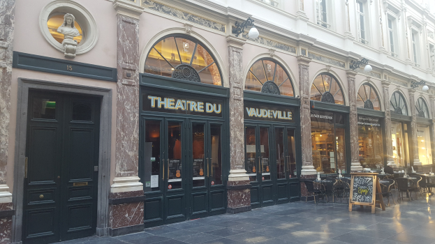 Theater du Vaudeville in Paris, Frankreich, zeigt eine Gebäudefassade mit Türen, Fenstern, beleuchteten Schildern, Tischen, Stühlen und einer Wandstatue.