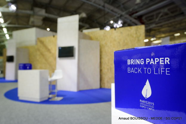 Messestände mit einem blauen und weißen "Bring Paper Back to Life"-Schild auf der rechten Seite, mit unscharfen Stühlen, Tischen, einem wandmontierten Fernseher und Deckenlampen im Hintergrund.